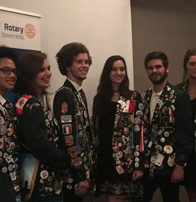 A group of five young people wearing dark green blazers completely covered in colorful international pins and flag patches.