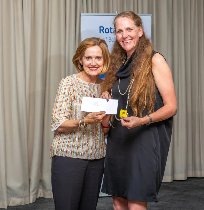 Two women standing in front of a Rotary banner; one in a patterned shirt hands an envelope to another in a black dress.