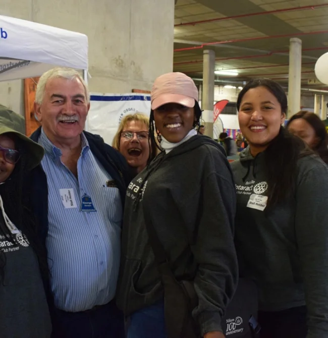 Four smiling people, including three young women in "Rotaract" hoodies and an older man, posing together at an indoor event.