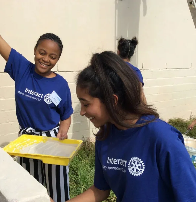 Two girls wearing blue "Interact Rotary Sponsored Club" t-shirts smiling while working on a community painting project.