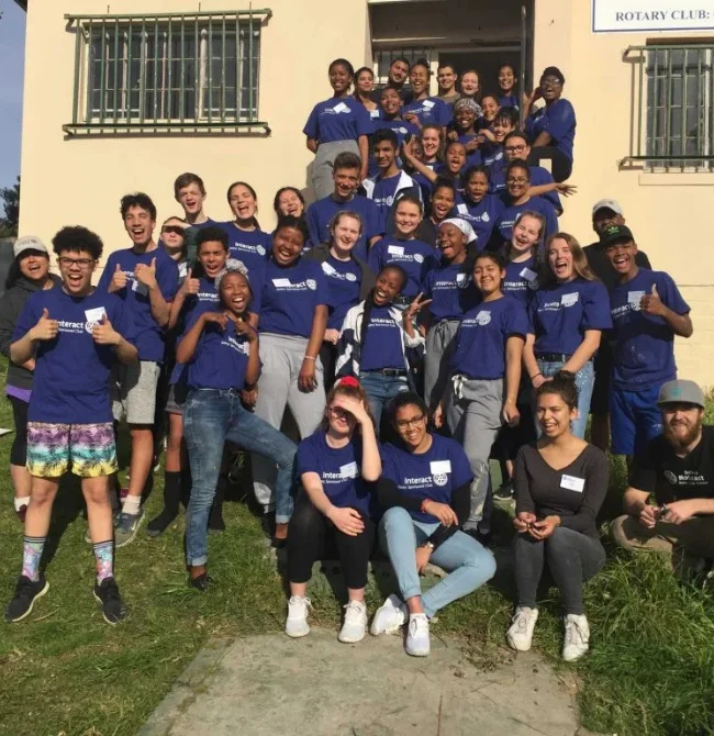 A large group of approximately 30 teenagers in matching blue Interact t-shirts posing and cheering on the steps of a yellow building.