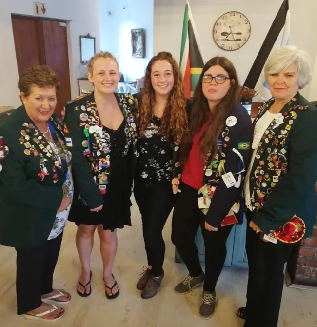 Five women standing in a line; three wear dark green blazers covered in pins, while two younger women in the center wear casual clothes.