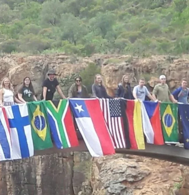 A group of youth exchange students standing on a bridge holding various international flags including South Africa, Brazil, USA, and Chile.