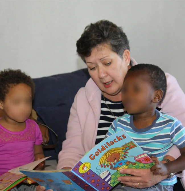 An adult woman reading a "Goldilocks" picture book to two young children sitting on her lap.