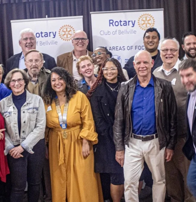 A diverse group of Rotary Club members standing together and smiling in front of "Rotary Club of Bellville" banners.